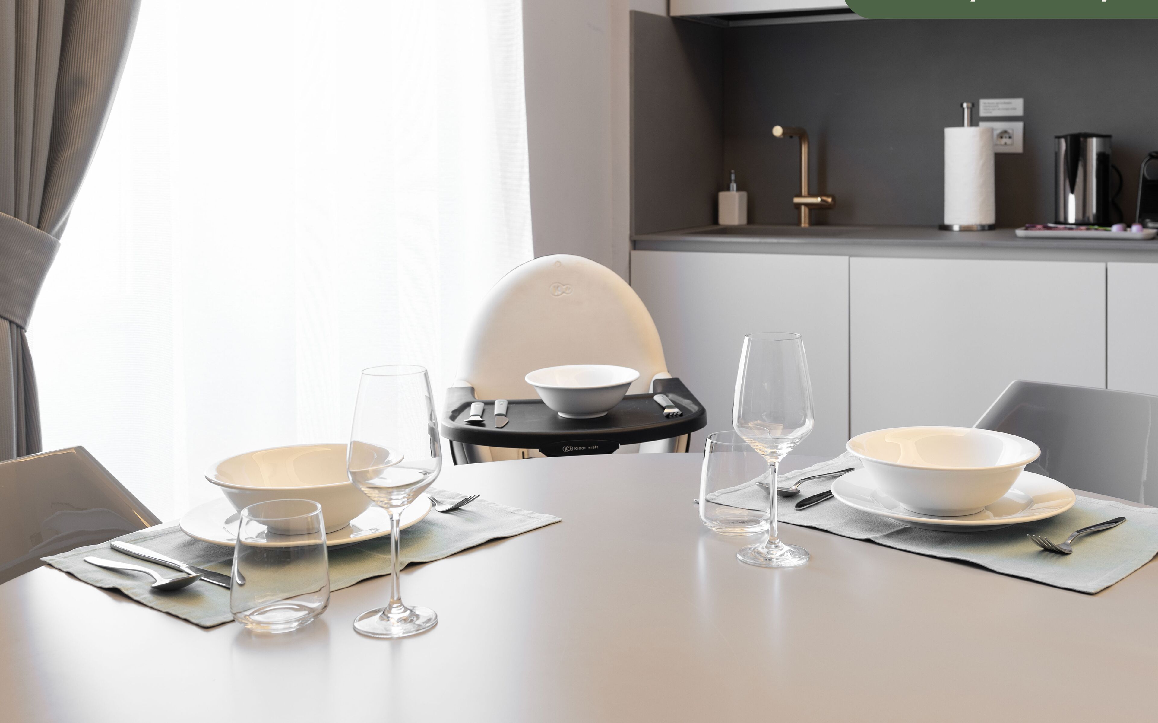 Una mesa de comedor dispuesta con fuentes, copas de vino y cubiertos, una trona, y una cocina con hervidor y utensilios.