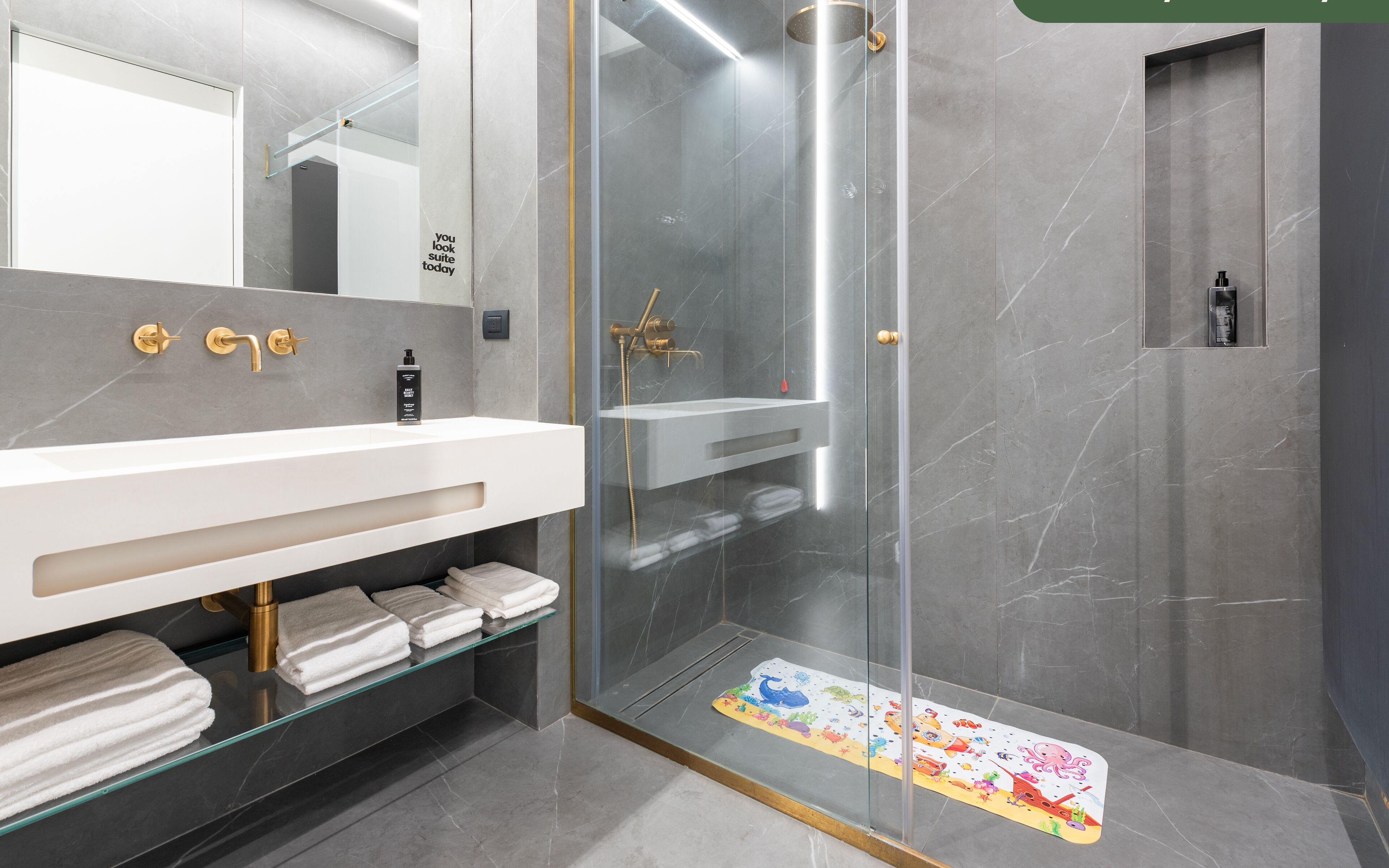 A modern bathroom with a glass-enclosed shower, a white vanity with gold fixtures, and a glass shelf holding towels.