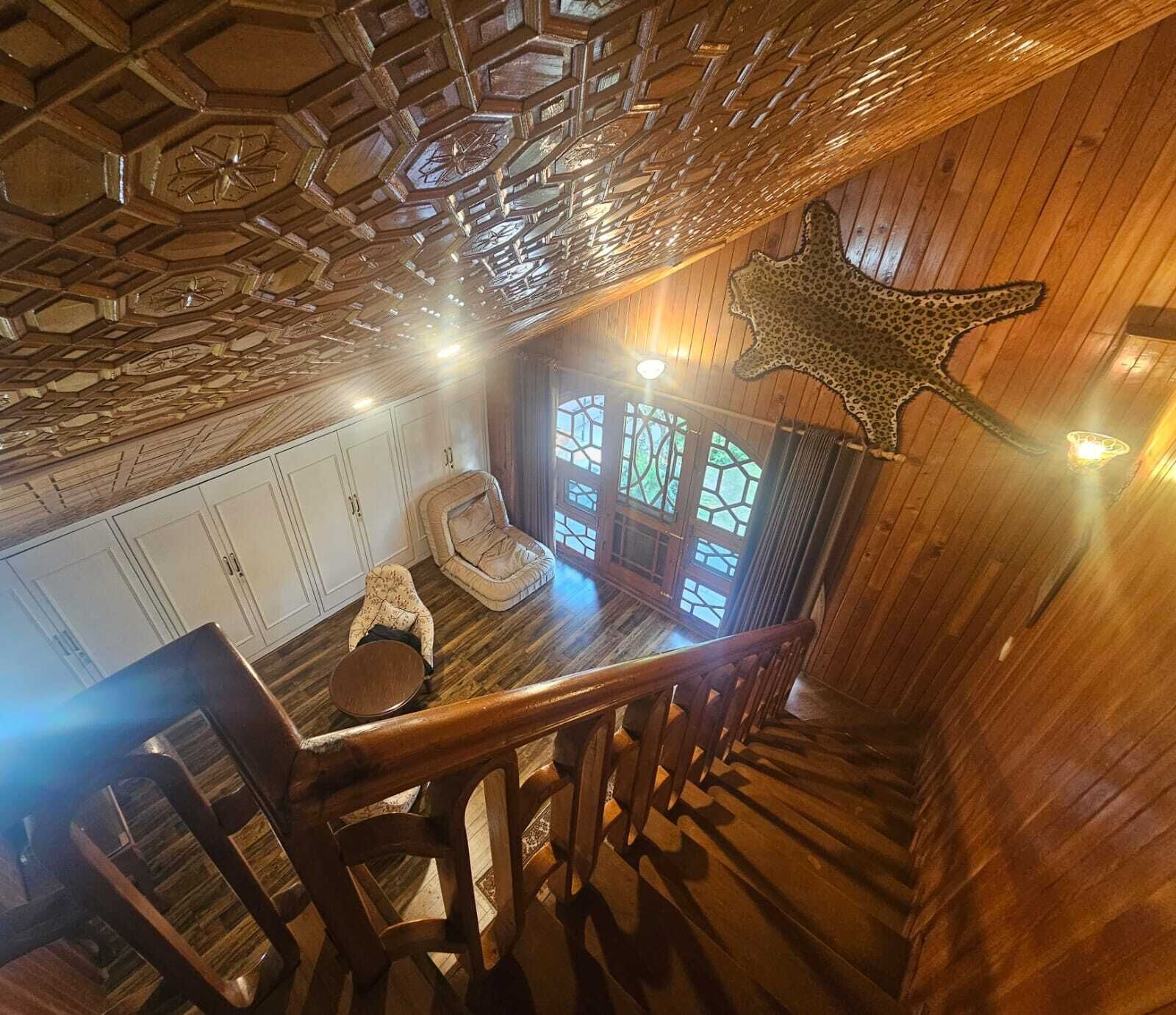 A wooden interior with a staircase, a leopard print wall hanging, and a room with a window and a sofa.
