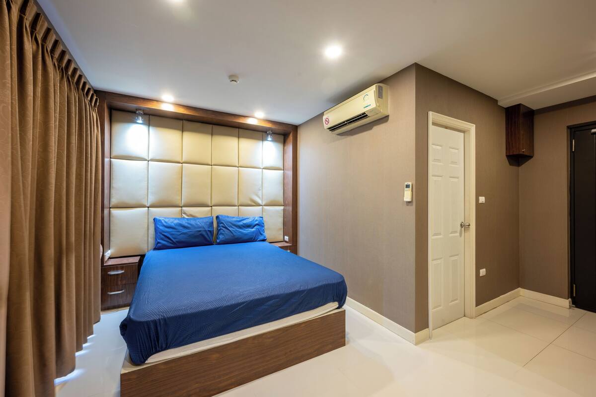 Un dormitorio con una cama grande, cabecera, mesita de noche, unidad de aire acondicionado y una puerta.