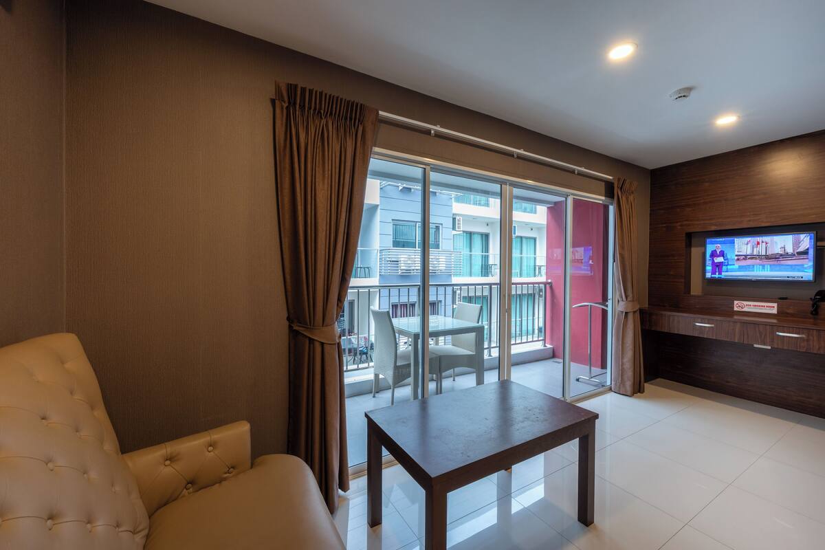 Habitación de hotel moderna con un amplio ventanal corredero que da a un balcón, un televisor de pantalla plana, una mesa de madera y un sofá confortable.