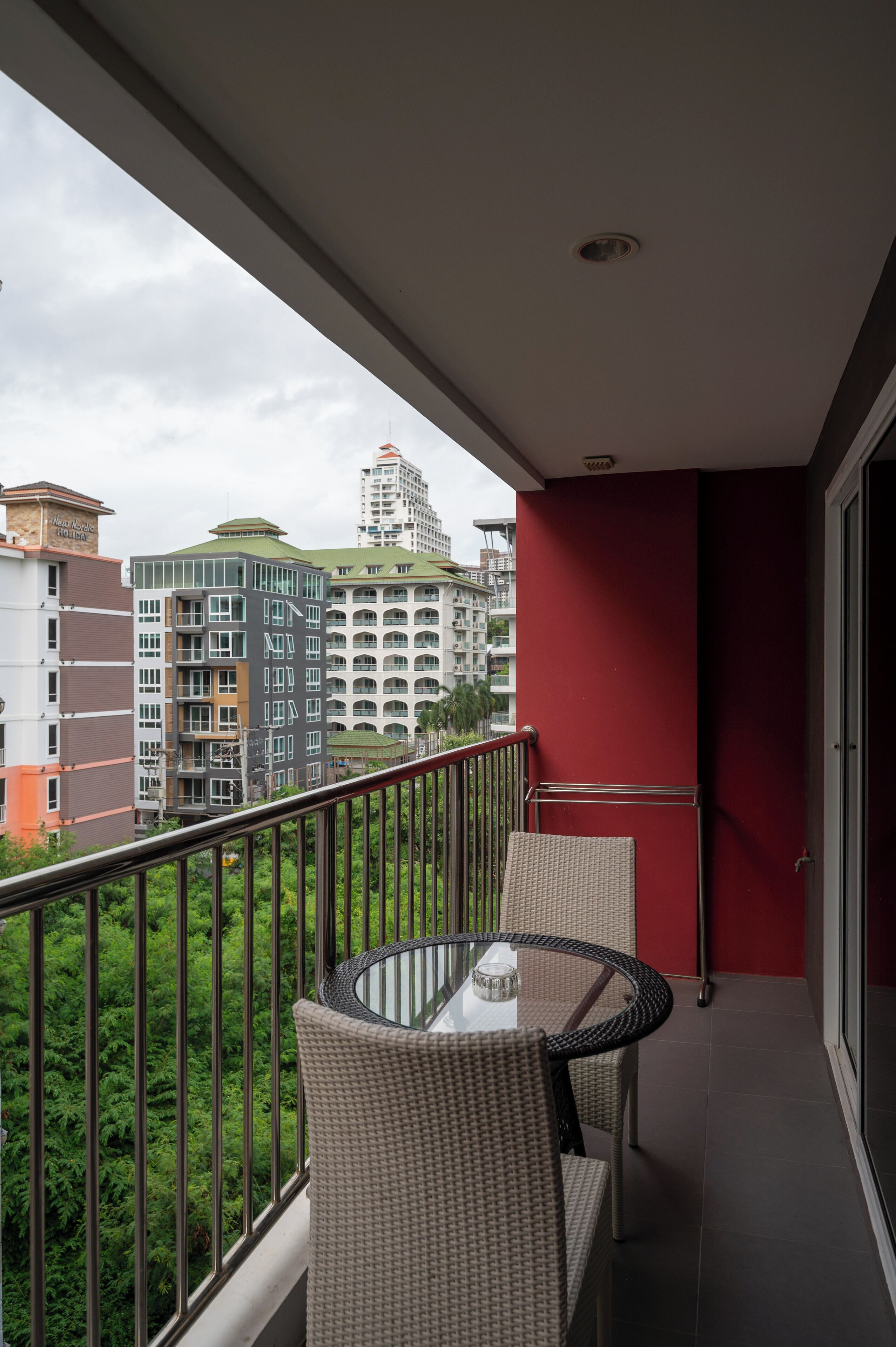 A balcony with a glass table and wicker chairs, overlooking a cityscape with various buildings and greenery.
