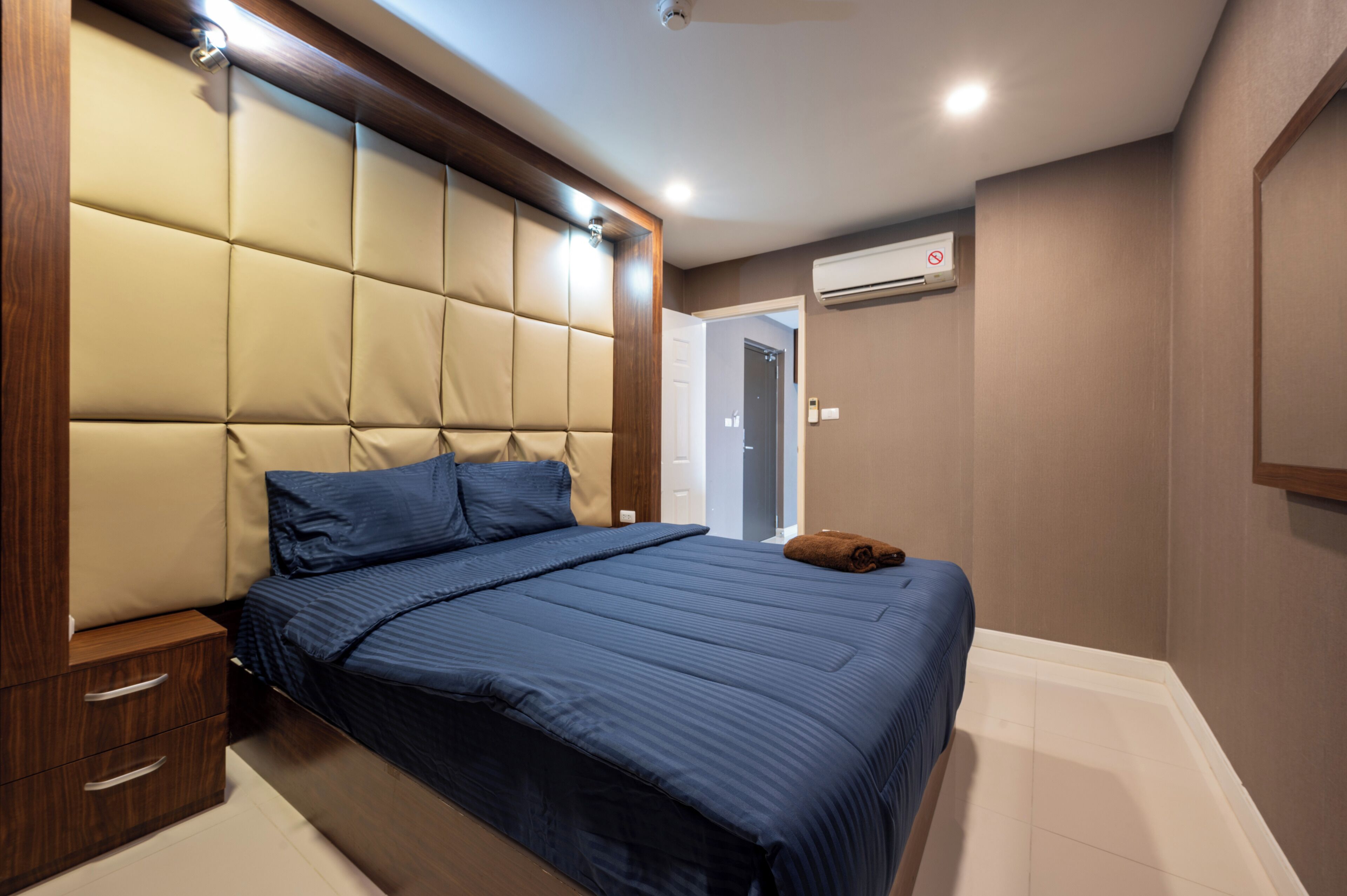 Un dormitorio con un cabecero capitonado, una cama con ropa de cama azul, una mesita de noche de madera y un aire acondicionado de pared.