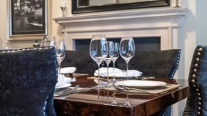 Une table à manger dressée avec des verres à vin, des assiettes et des couverts, avec une cheminée et un tableau encadré en arrière-plan.