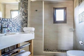 A bathroom with a stone wall, a shower, a toilet, and a sink with toiletries. - Villa Pomegranate Tree (Kefalonia)