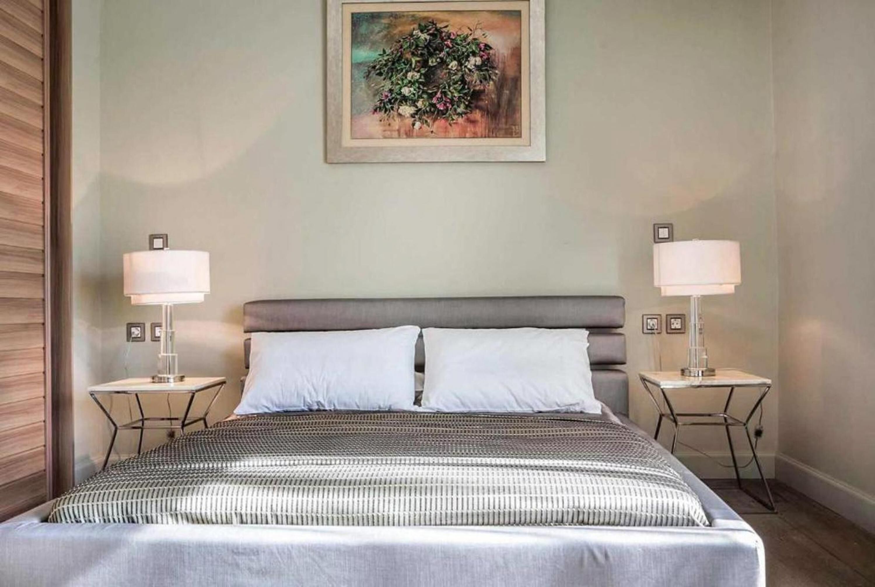 A hotel room with a large bed, two bedside tables with lamps, a framed painting on the wall, and a wooden panel on the left side.