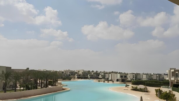 A resort with a large swimming pool, palm trees, and modern buildings in the background. - Sand Castle, Private Villa in Azha, Ain Sokhna (Ataqah)