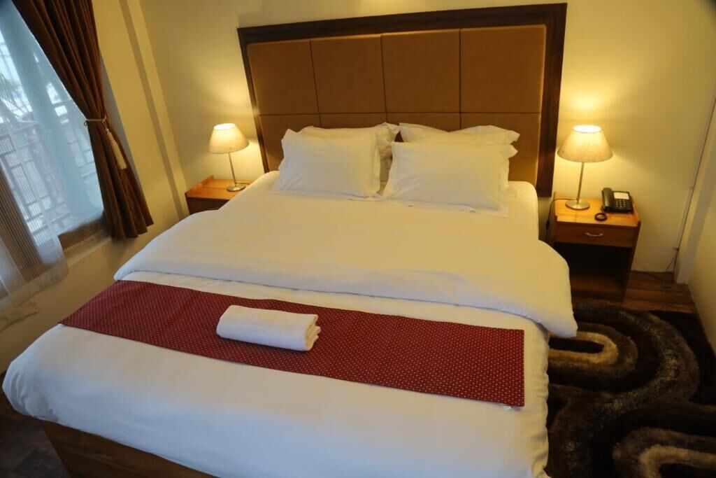 Une chambre d’hôtel avec un grand lit, deux tables de chevet avec des lampes, une fenêtre avec des rideaux et un tapis rouge sur le lit.