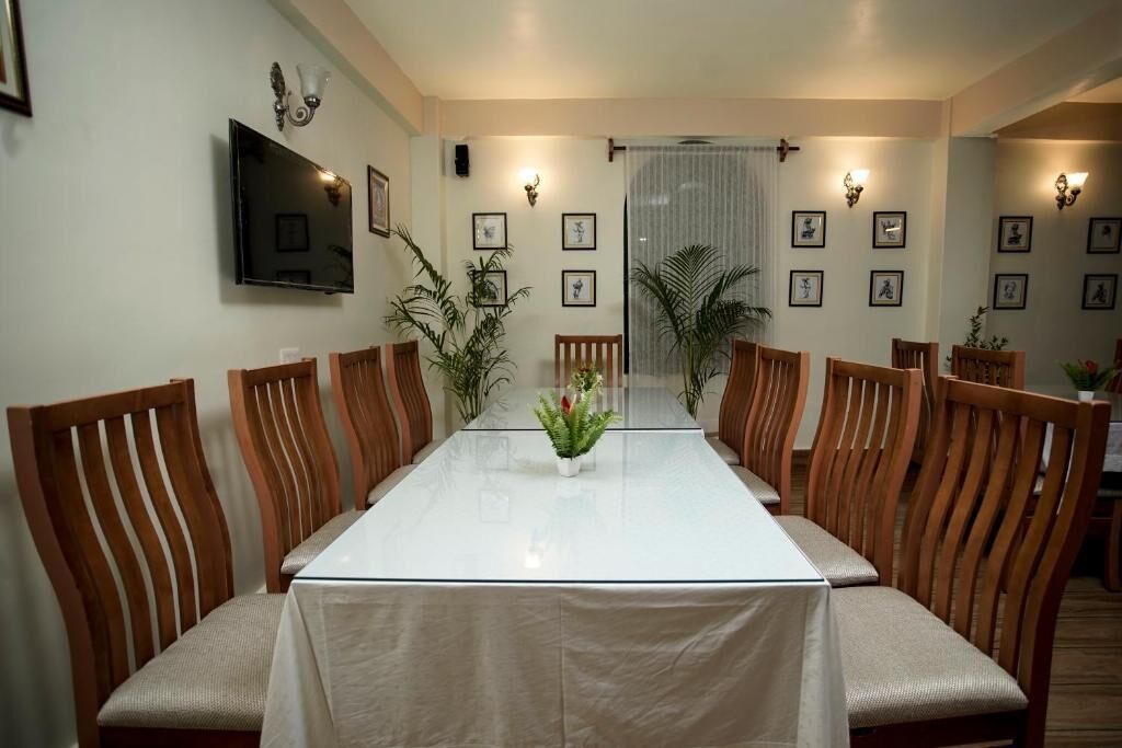 Une salle de conférence avec une longue table, des chaises en bois, des plantes en pot, des tableaux encadrés accrochés aux murs et un téléviseur fixé au mur.