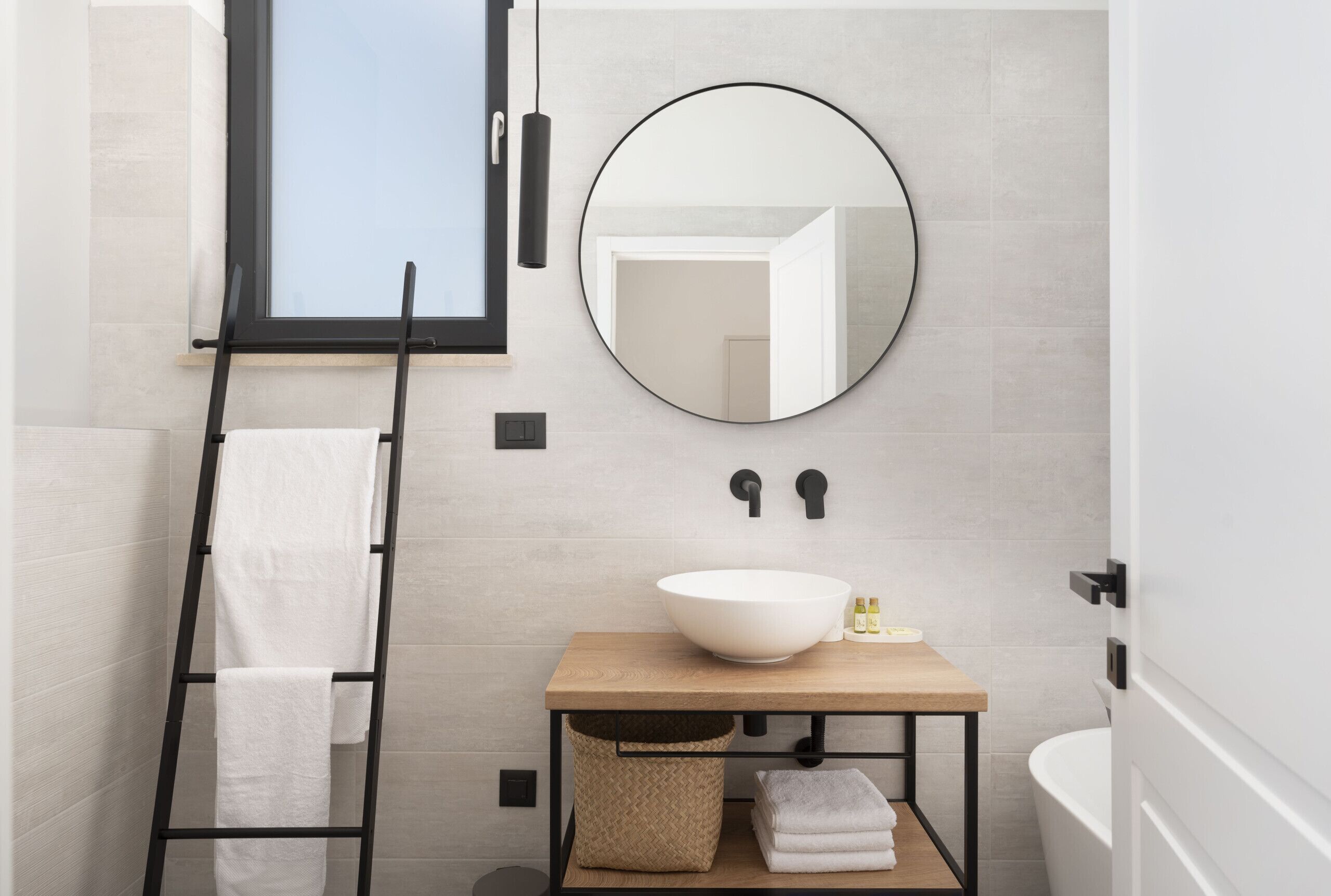 A bathroom with a wooden vanity, a round mirror, a towel rack, and a window.