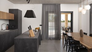 Eine moderne Küche mit einer zentralen Kochinsel, einem Essbereich mit Holztisch und -stühlen sowie einem Ausblick durch ein großes Fenster mit Vorhängen.