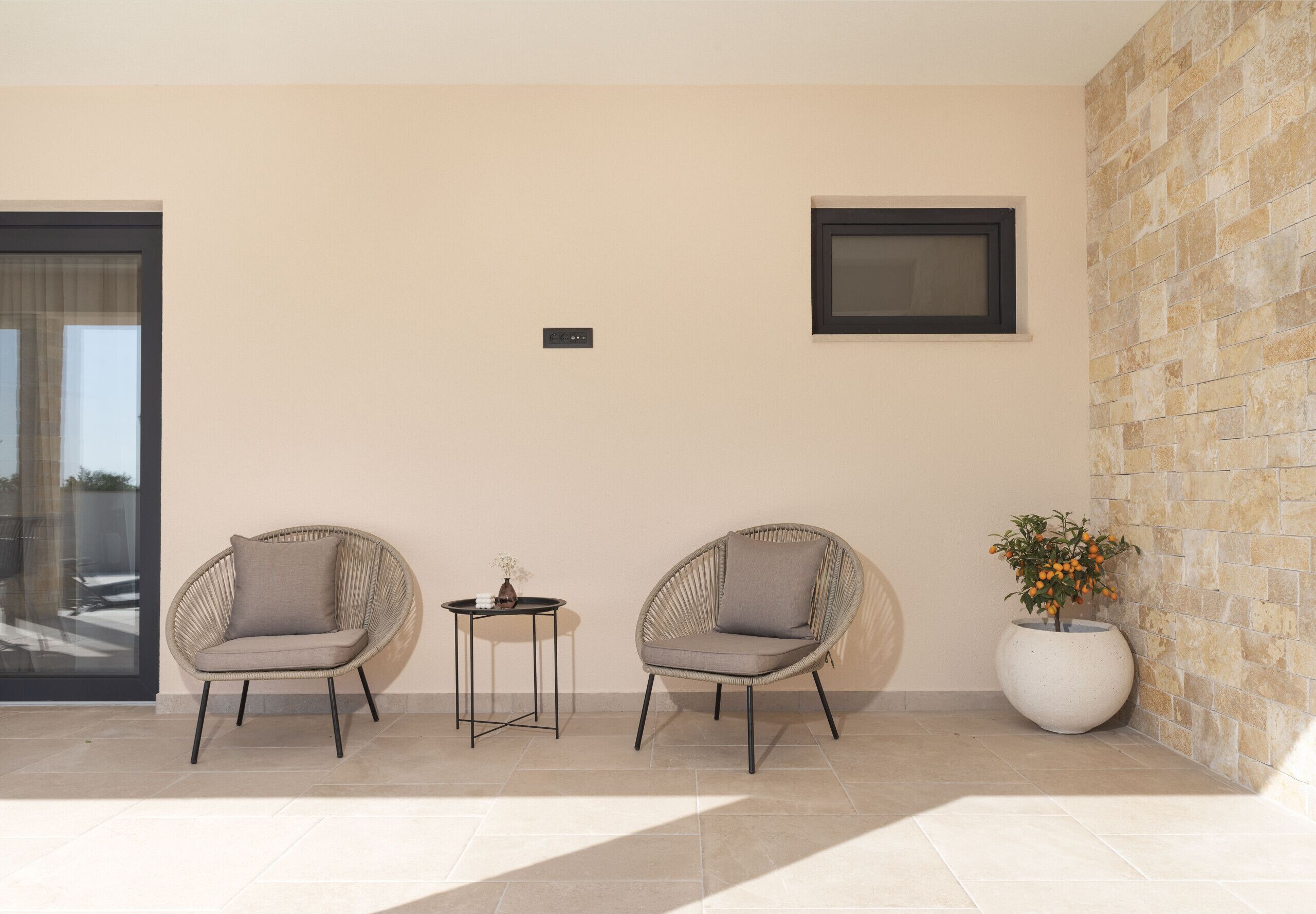 A modern outdoor seating area with two wicker chairs and a small table, a potted plant, and a framed picture on the wall.