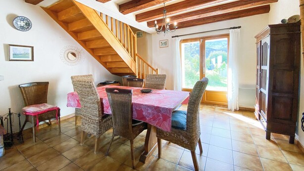 Un coin repas avec un escalier en bois, des chaises en osier, une table recouverte d’une nappe rose et une vue dégagée sur l’extérieur grâce à une grande fenêtre.