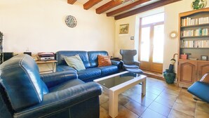 Un salon avec un canapé en cuir bleu, une table basse en verre, des poutres en bois au plafond, une bibliothèque et une porte donnant sur l’extérieur.