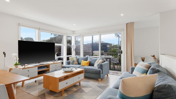 A modern living room with a flat-screen TV, a wooden coffee table, a grey sofa, and a dining area with a table and chairs.