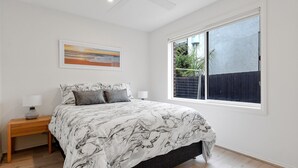 A bedroom with a large bed, a nightstand, a lamp, and a window with a view of a fence and greenery.