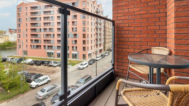 Un balcone con sedie in vimini e un tavolo di vetro, con vista su un parcheggio e un edificio a più piani.