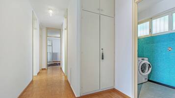 A hallway with wooden flooring, a white wardrobe, and a washing machine in a bathroom with a blue-tiled wall.