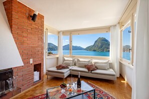 A modern living room with a brick fireplace, a sofa set, a glass coffee table with drinks, and a scenic mountain and lake view. - Casa Serenella Splendid Lake View (Lugano)