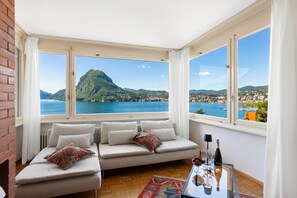 A room with a sofa, a glass table with a bottle and glasses, and large windows with a mountain and sea view. - Casa Serenella Splendid Lake View (Lugano)