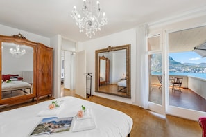 A bedroom with a large bed, a wooden wardrobe, a chandelier, a large mirror, and a balcony with a view. - Casa Serenella Splendid Lake View (Lugano)