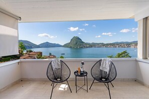 A balcony with two chairs and a small table, overlooking a lake with mountains and a town. - Casa Serenella Splendid Lake View (Lugano)