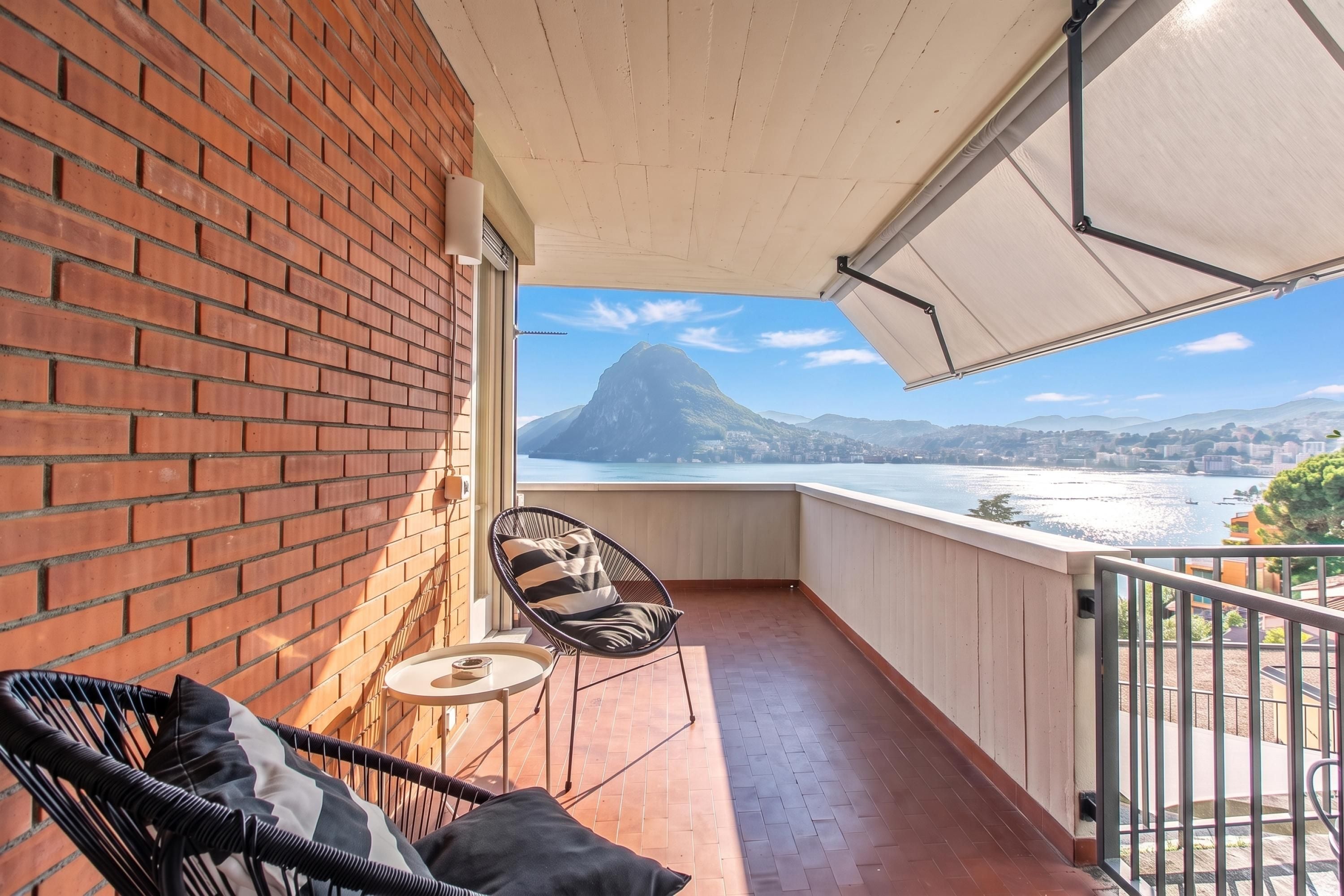 Een balkon met een bakstenen muur, een stoel, een klein tafeltje en uitzicht op een berg en water.
