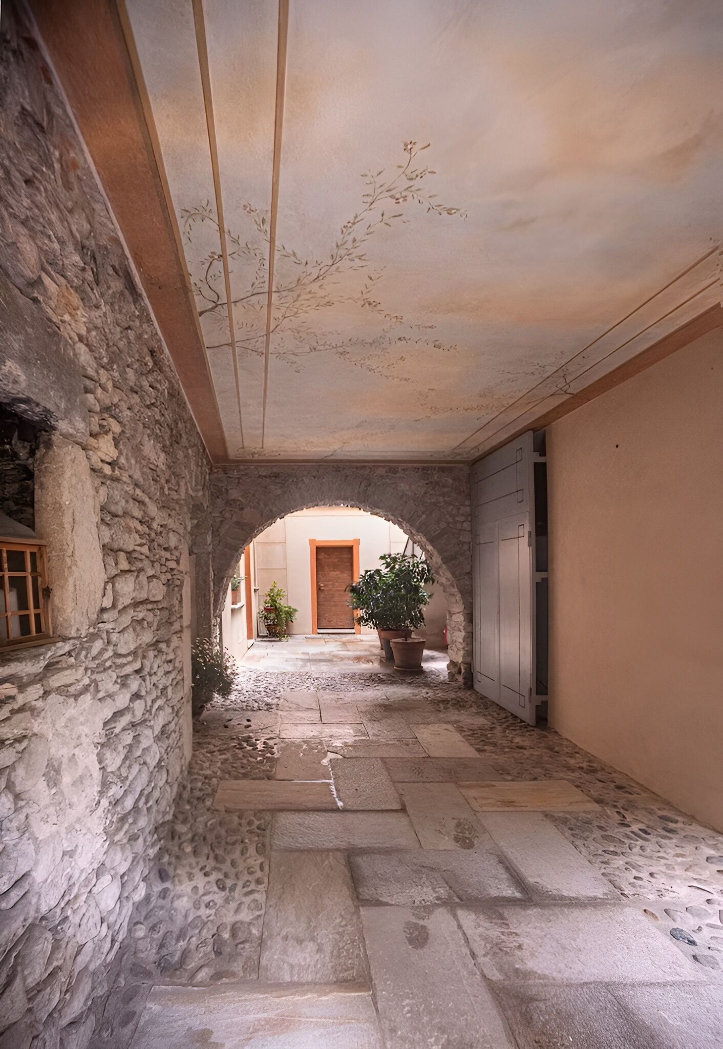 Un corridoio con pareti in pietra, un arco, piante in vaso e una porta in lontananza.
