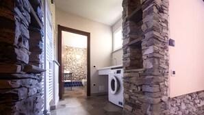 A laundry area with a washing machine, a stone wall, and a view into a room with a stone wall and a blue tiled floor. - Apartment in historic building - blue (Bedero)