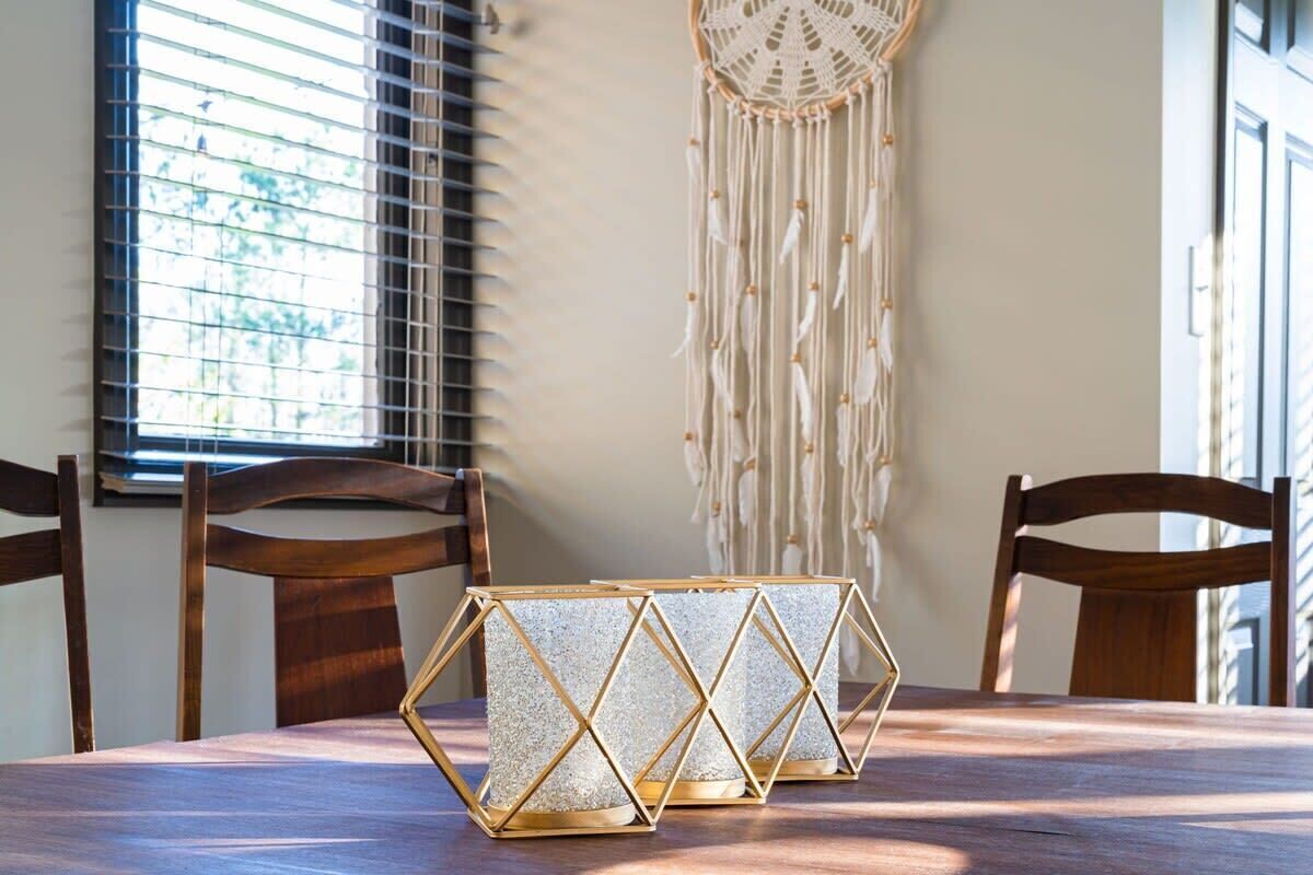 A dining table with a geometric patterned table runner, wooden chairs, a macrame wall hanging, and a window with blinds.
