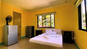 Una camera d'albergo con un letto, un frigorifero, una ventola, una cassettiera e una finestra vista verde.