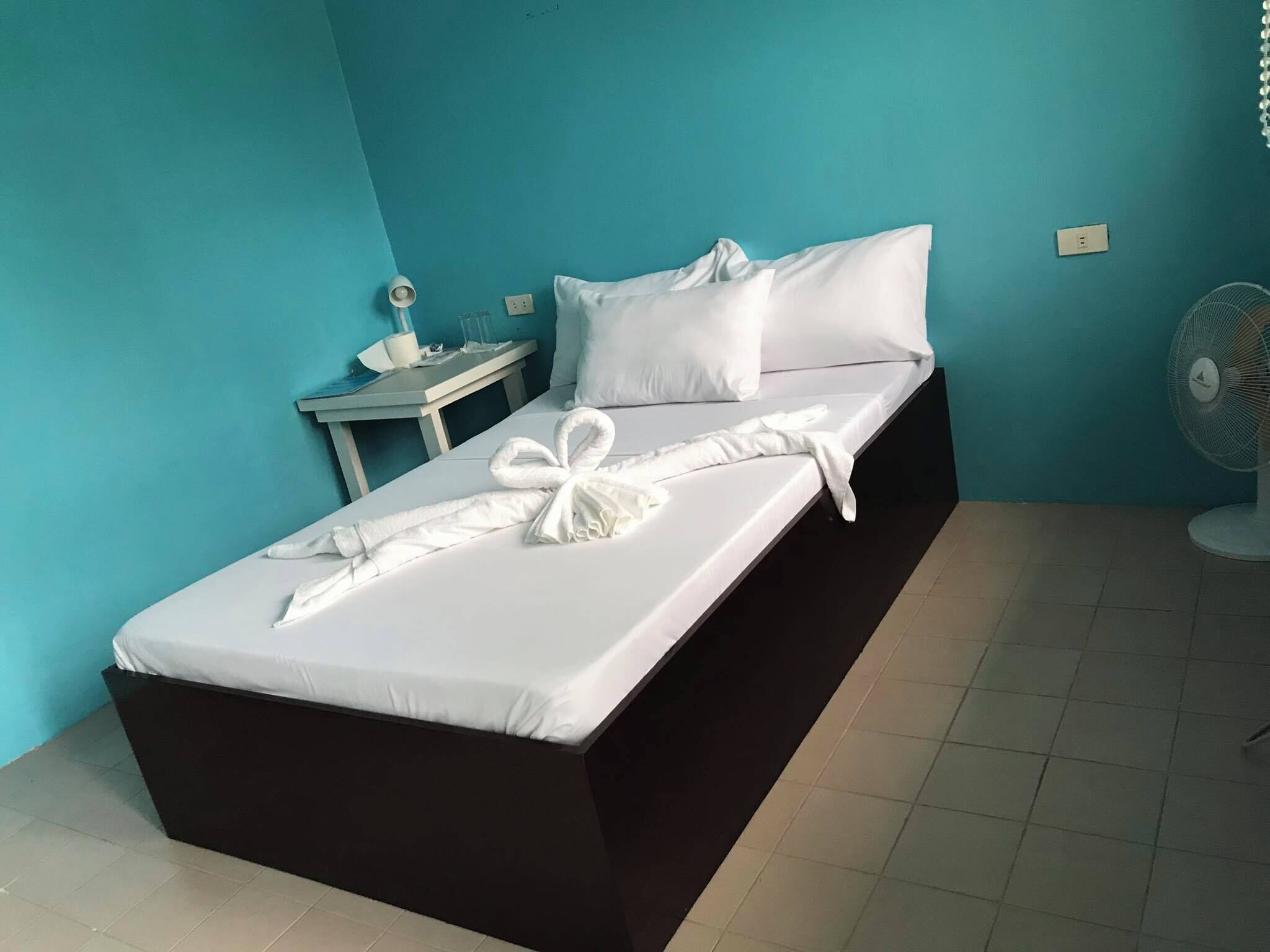 Una cama bien hecha con sábanas blancas y una toalla, una mesita de noche con lámpara y un ventilador de pie.