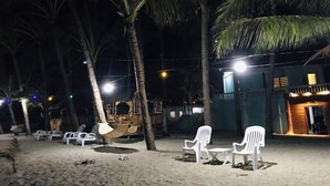 Area sulla spiaggia con sedie di plastica bianca e un'amaca, palme e un edificio con un cartello che riporta la scritta 'ENTRANCE'.