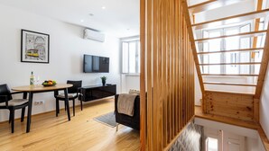 Un saló modern amb escala de fusta, zona de menjador amb taula i cadires, sofà negre, televisió penjada a la paret i finestra que deixa entrar llum natural.