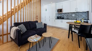 Un saló modern amb un sofà negre, una escala de fusta i una zona de cuina amb armaris blancs i un microones.