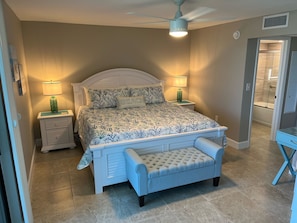 A bedroom with a bed, nightstands, a bench, a ceiling fan, and a bathroom visible through an open door.