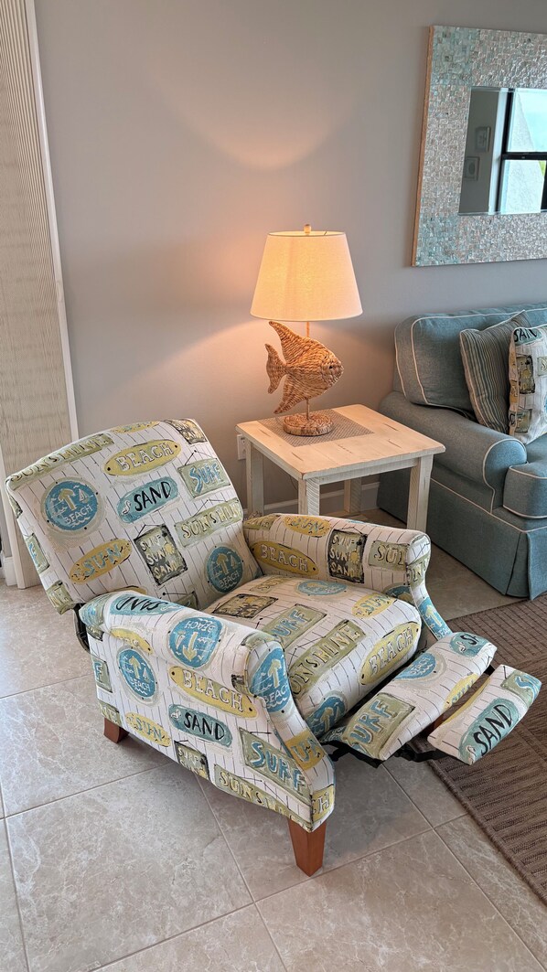 Un fauteuil sur le thème de la plage, avec une lampe en forme de poisson tressé placée à côté, une petite table en bois et un canapé avec des coussins à rayures.