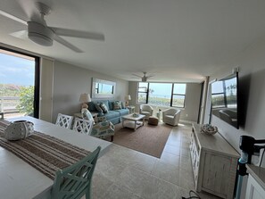 A spacious living room with a large sofa, two armchairs, a coffee table, and a dining area with a table and chairs. There is a ceiling fan, large windows with a view, and a flat-screen TV mounted on the wall.
