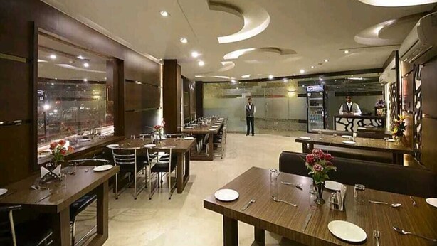 A modern restaurant with a long counter and multiple tables, a waiter in the background, and a view of the city through the windows. - Hotel Mani Ram Palace (Agra)