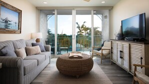 A living room with a sofa, ottoman, TV, and a balcony with a view. - Seaside Serenity-2BD 2BA condo w/ pool, parking, tennis courts, near beaches (Key West)