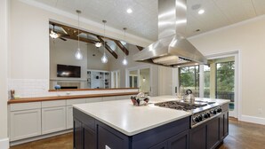 Une cuisine spacieuse dotée d’un grand îlot central, de poutres en bois, d’une hotte en acier inoxydable et de luminaires suspendus.