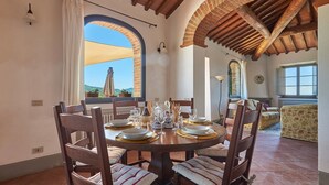 A dining area with a round table set for six, surrounded by wooden chairs with striped cushions. There are two windows with curtains, a brick wall, and a view of greenery outside. - Casa Luce, San Donato in Perano, Gaiole in Chianti, Siena and Chianti (Gaiole in Chianti)