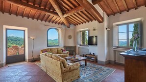 A spacious living room with a high ceiling, a fireplace, a television, and a large window overlooking a scenic landscape. - Casa Luce, San Donato in Perano, Gaiole in Chianti, Siena and Chianti (Gaiole in Chianti)