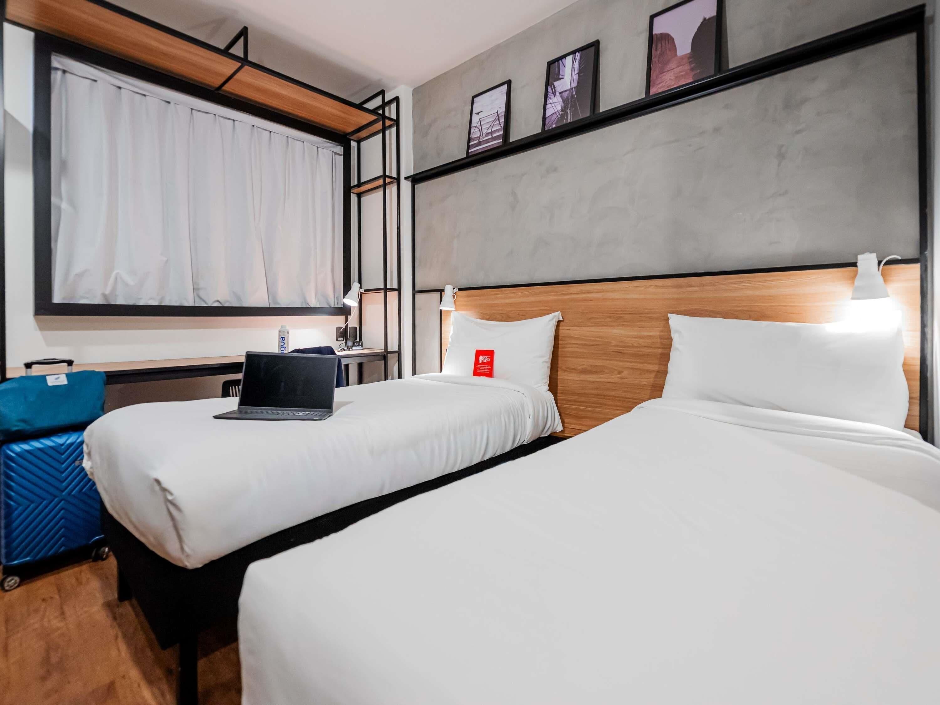 A hotel room with two beds, a desk, a laptop, a suitcase, and a window with curtains.