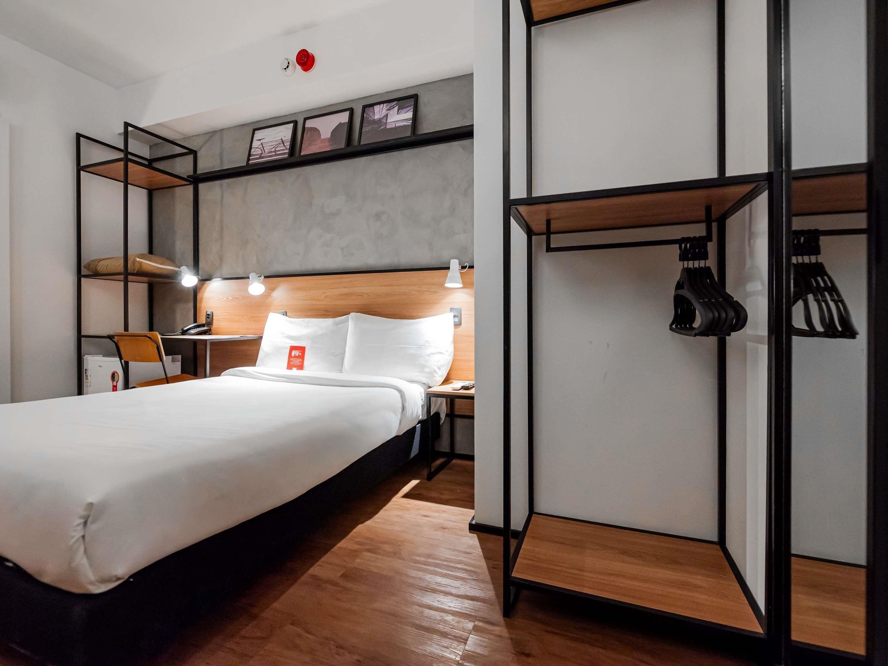 A hotel room with a bed, a desk, a chair, a wardrobe, and a shoe rack.