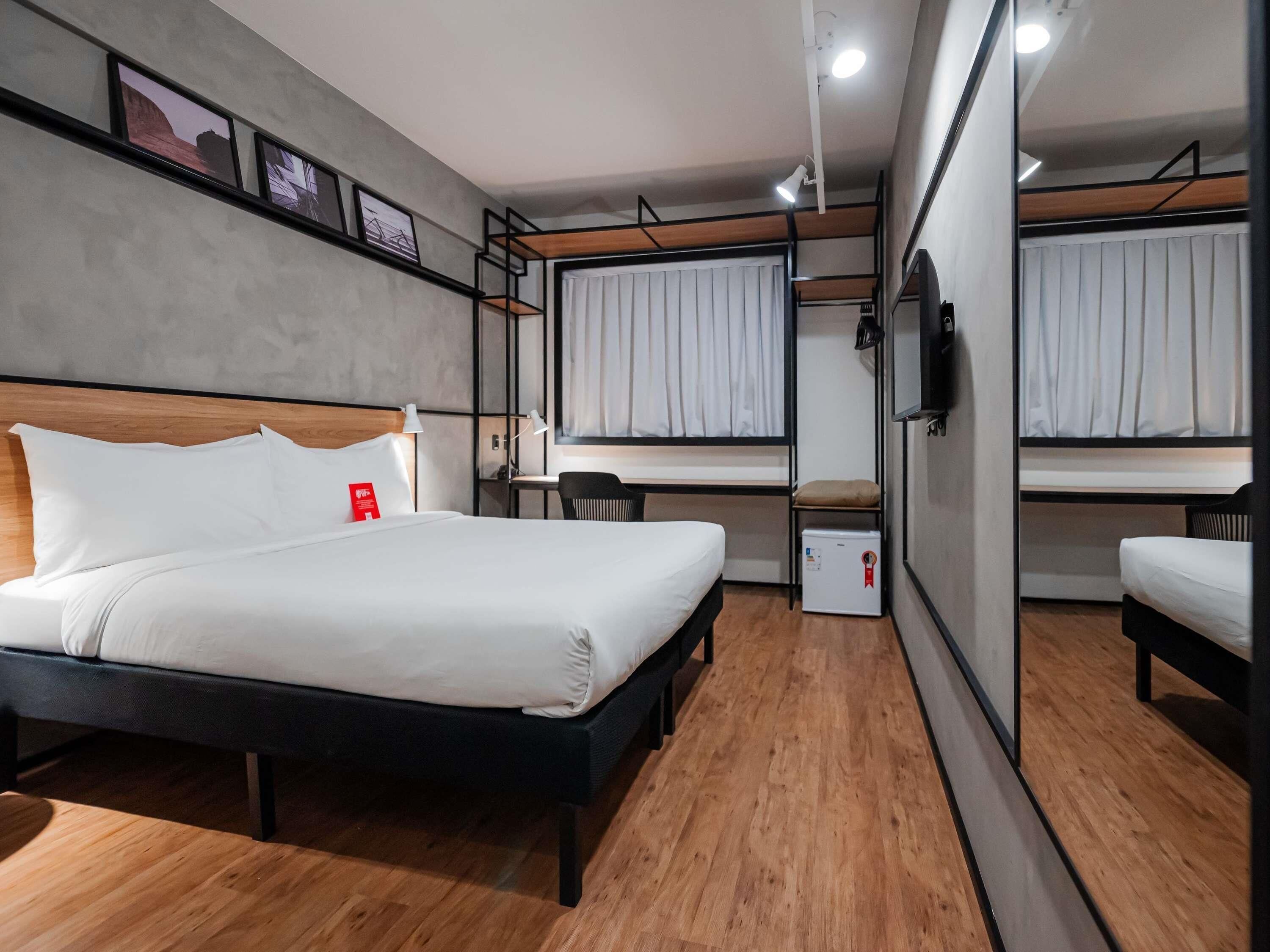 A hotel room with a bed, a desk, a chair, a small refrigerator, and a mirror.