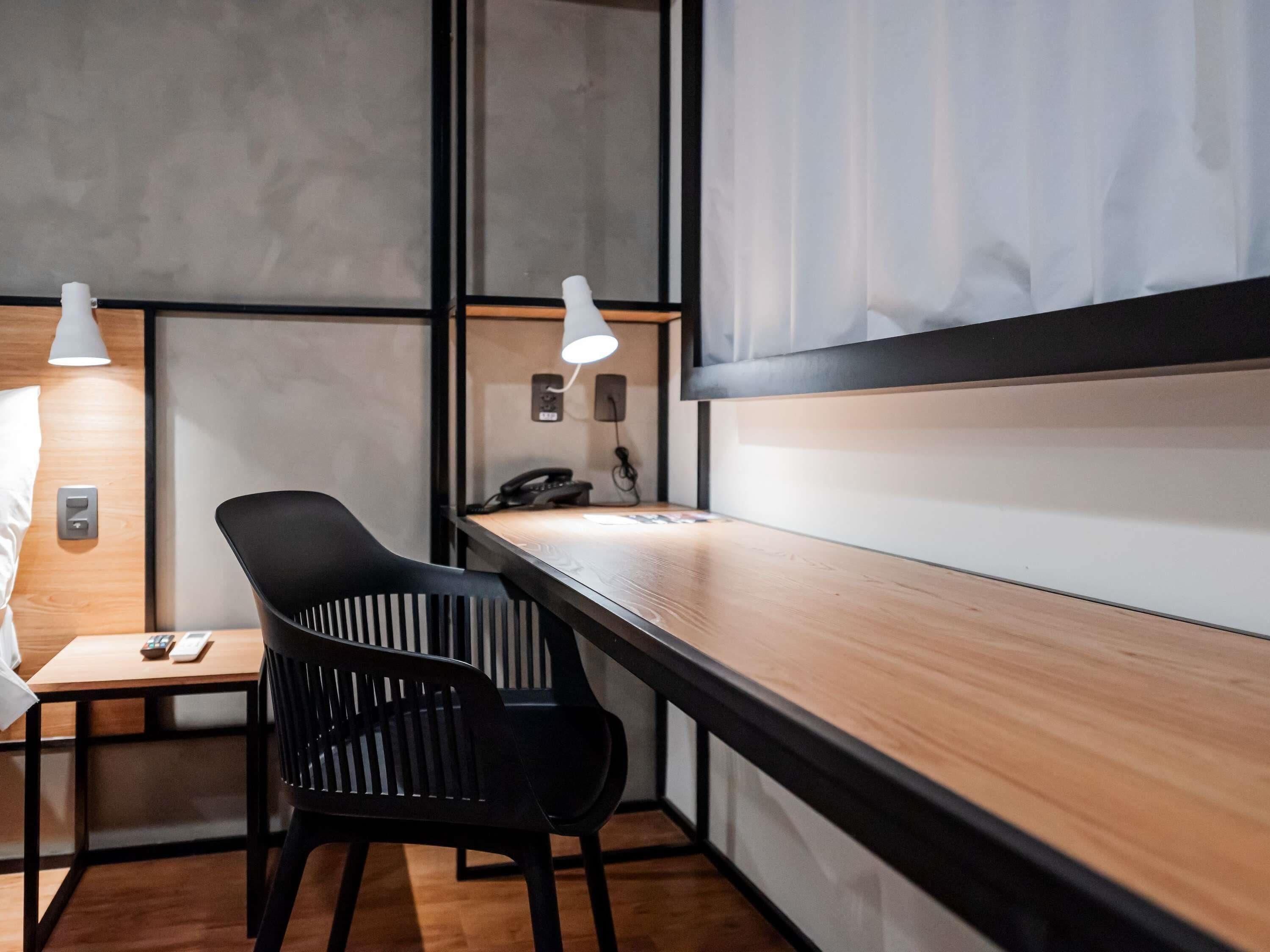 Una habitación de hotel moderna con un escritorio de madera, una silla negra, una mesita de noche con lámpara y un teléfono en la pared.
