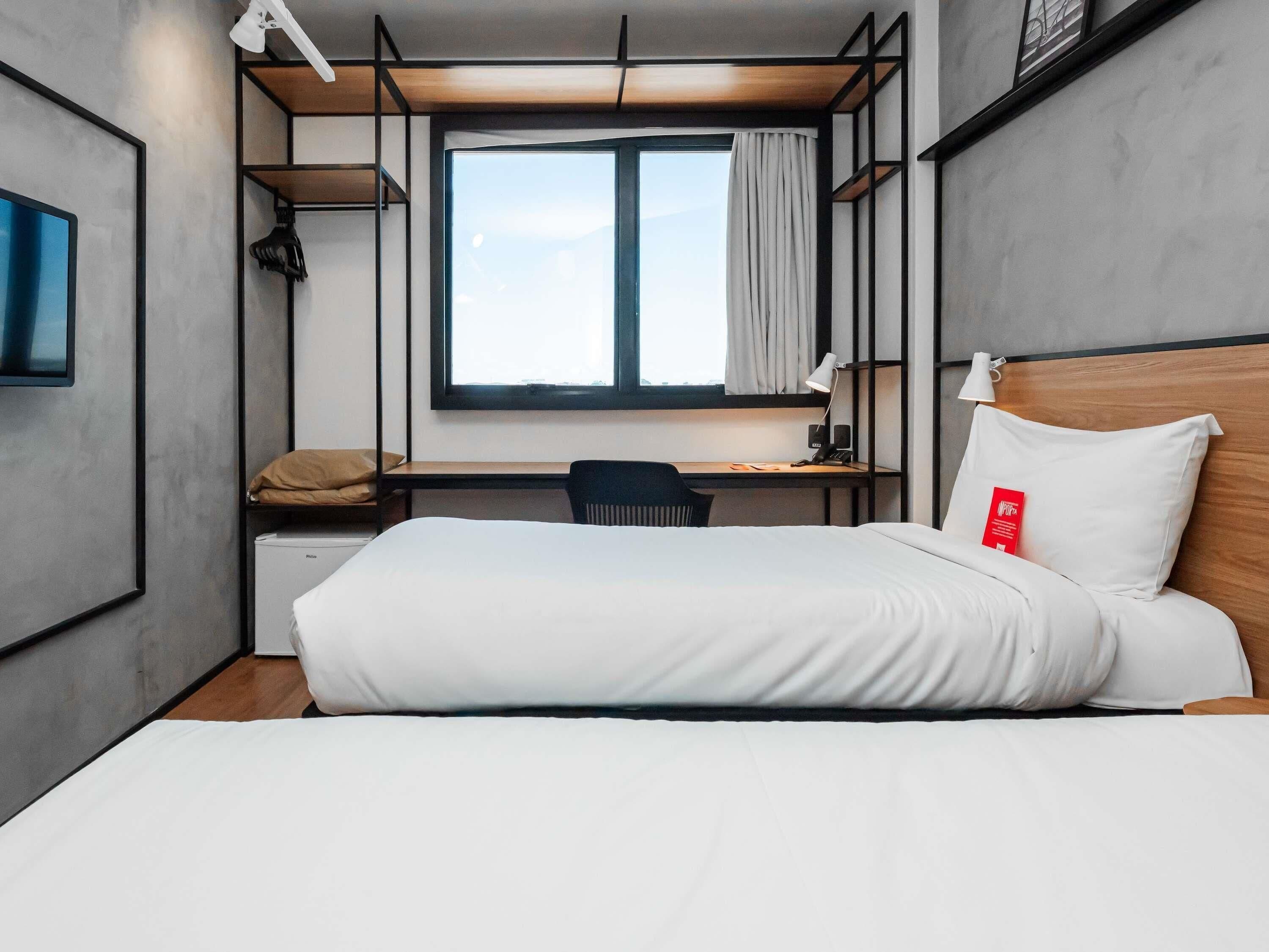Een moderne hotelkamer met een groot bed, een bureau en een stoel. Er is een raam met gordijnen, een lamp op het bureau en een rood voorwerp op het bed.