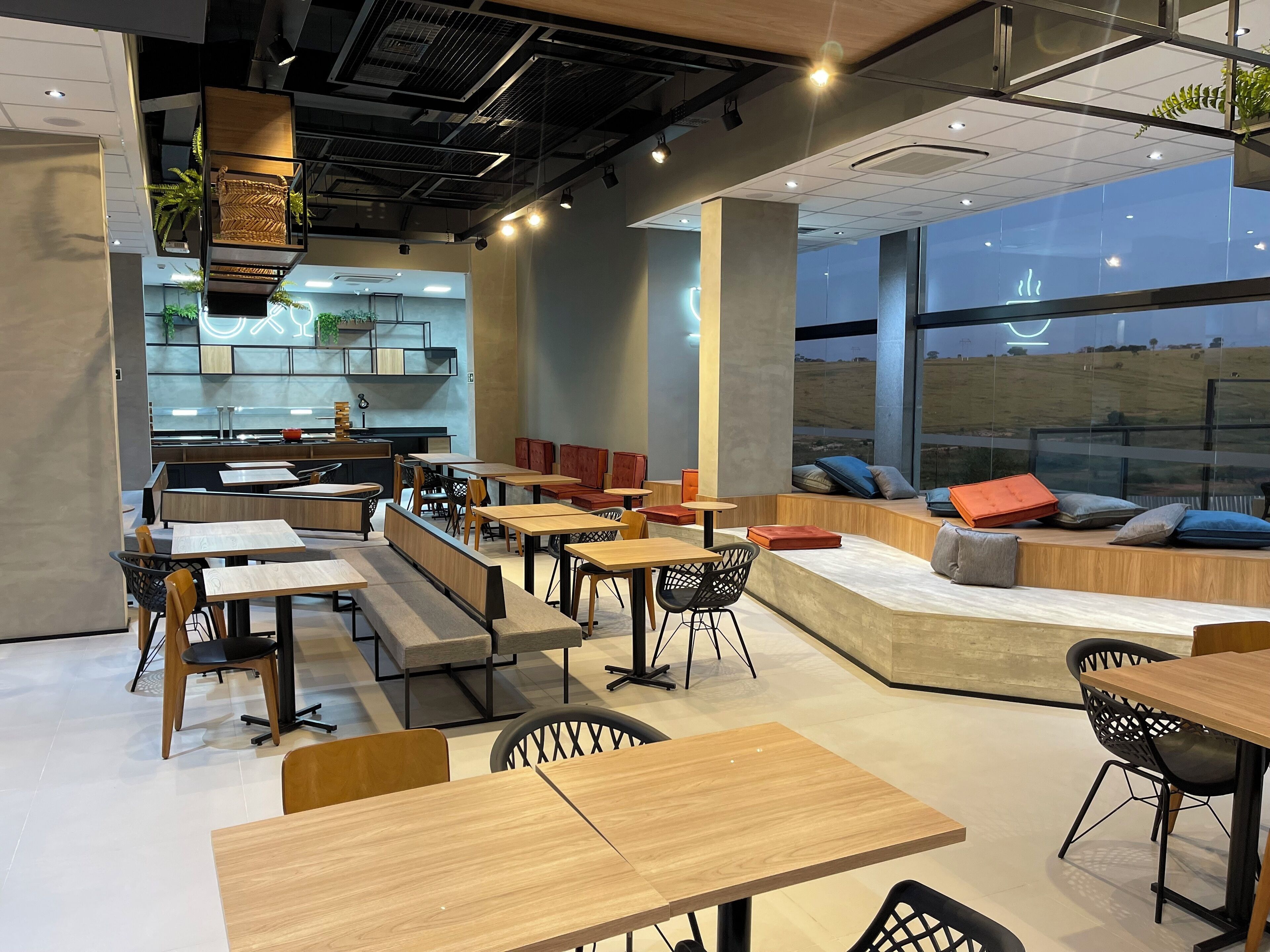 Un café moderno con mesas de madera y sillas negras, un área de estar con cojines, y ventanas grandes que ofrecen vistas al campo.