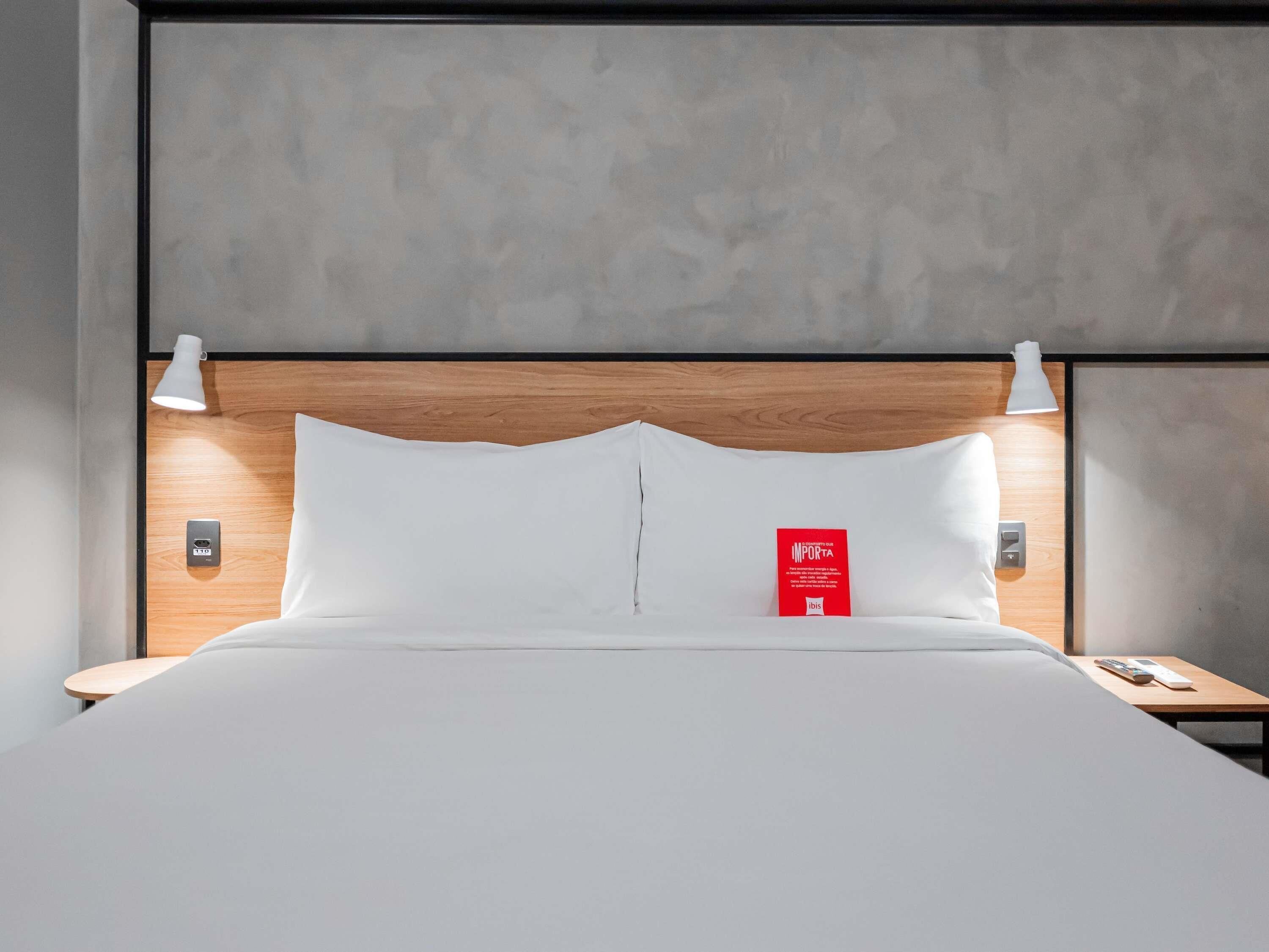 Cama bem arrumada com cabeceira de madeira, duas luminárias de apoio e um letreiro vermelho na cabeceira.